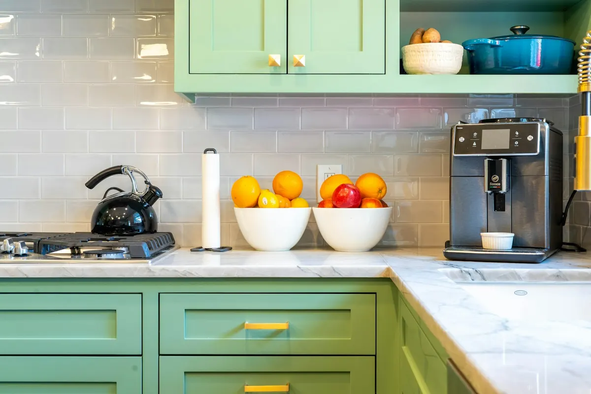 A kitchen corner with light green shaker cabinets, white marble-look counters, a white subway-tile backsplash and gold pulls