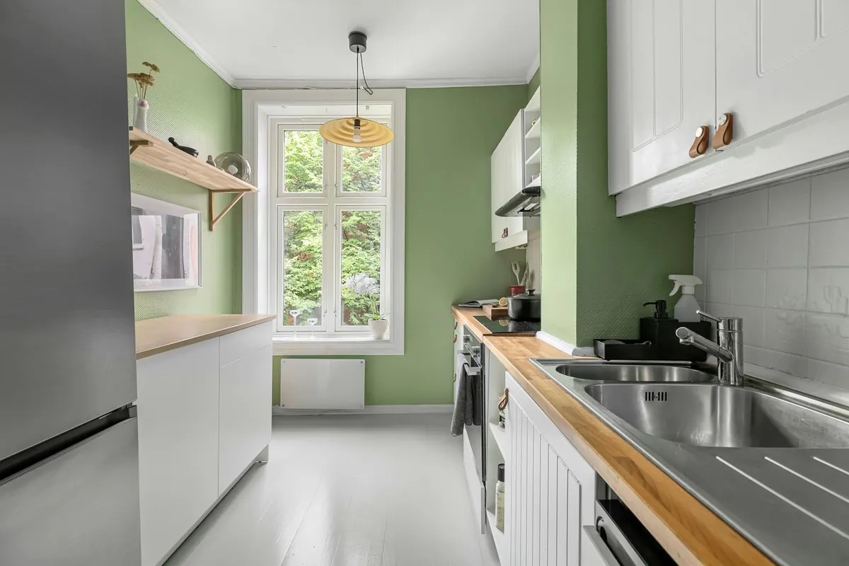 A narrow kitchen with sage-green walls, white upper cabinets, a butcher-block counter and a window at the far end