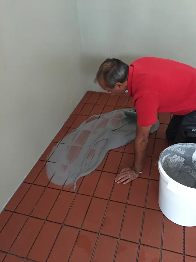 A tiler pressing grout into the joints of a red tile floor with a rubber float