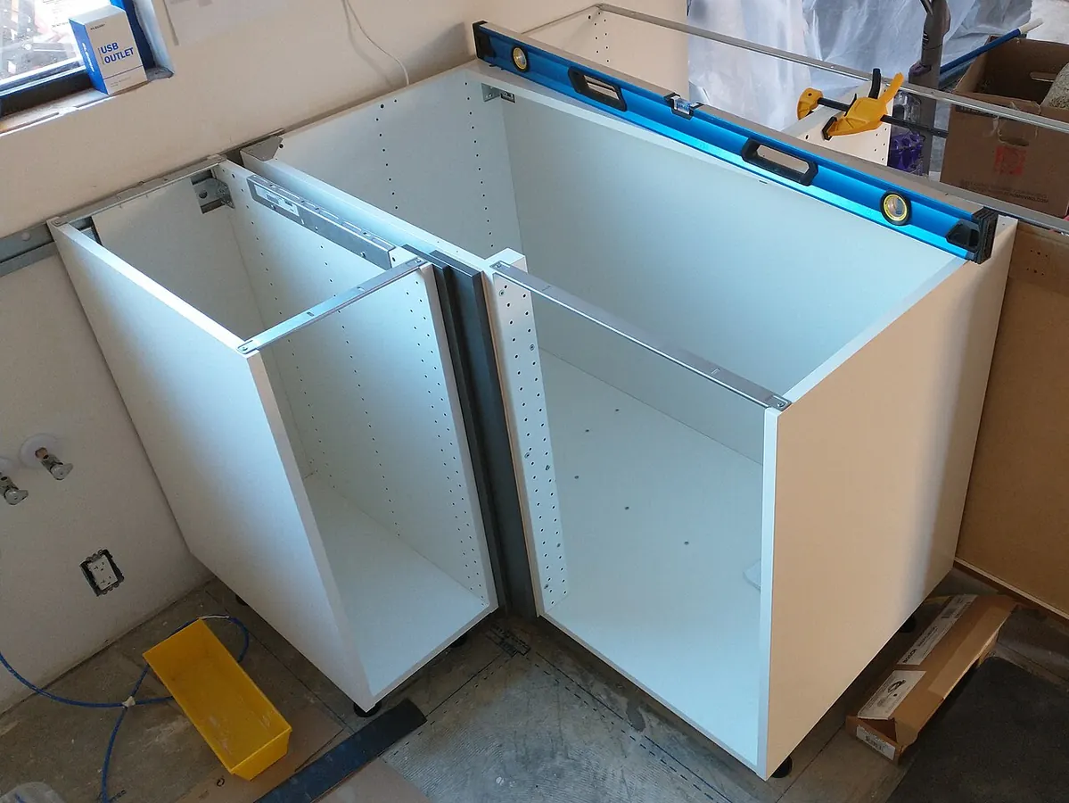Two assembled white flat-pack base cabinets, including a corner unit, set in a stripped kitchen with a level and clamps on top