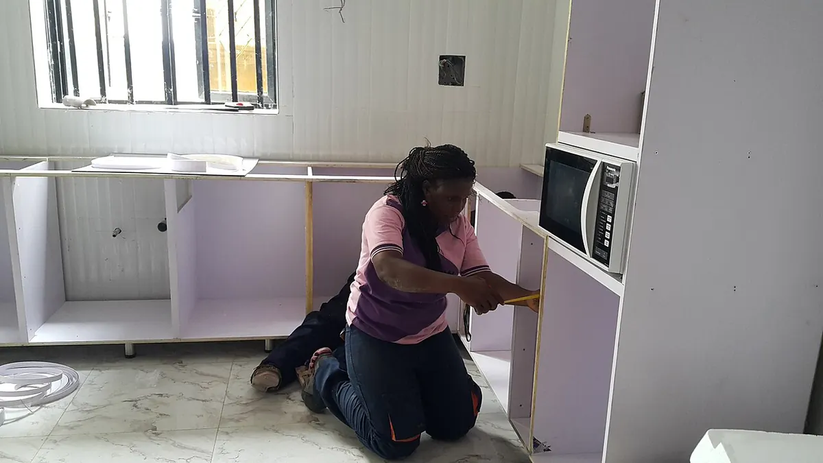 A worker kneeling on a marble-look floor fitting a row of new base cabinets along a kitchen wall, with an upper cabinet and microwave beside her