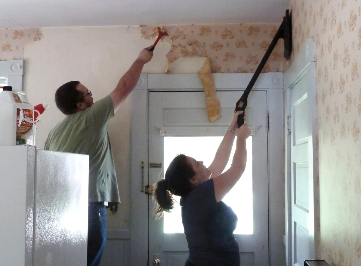 Two people stripping old floral wallpaper above a kitchen door, one with a hand scraper and one with a long-handled tool