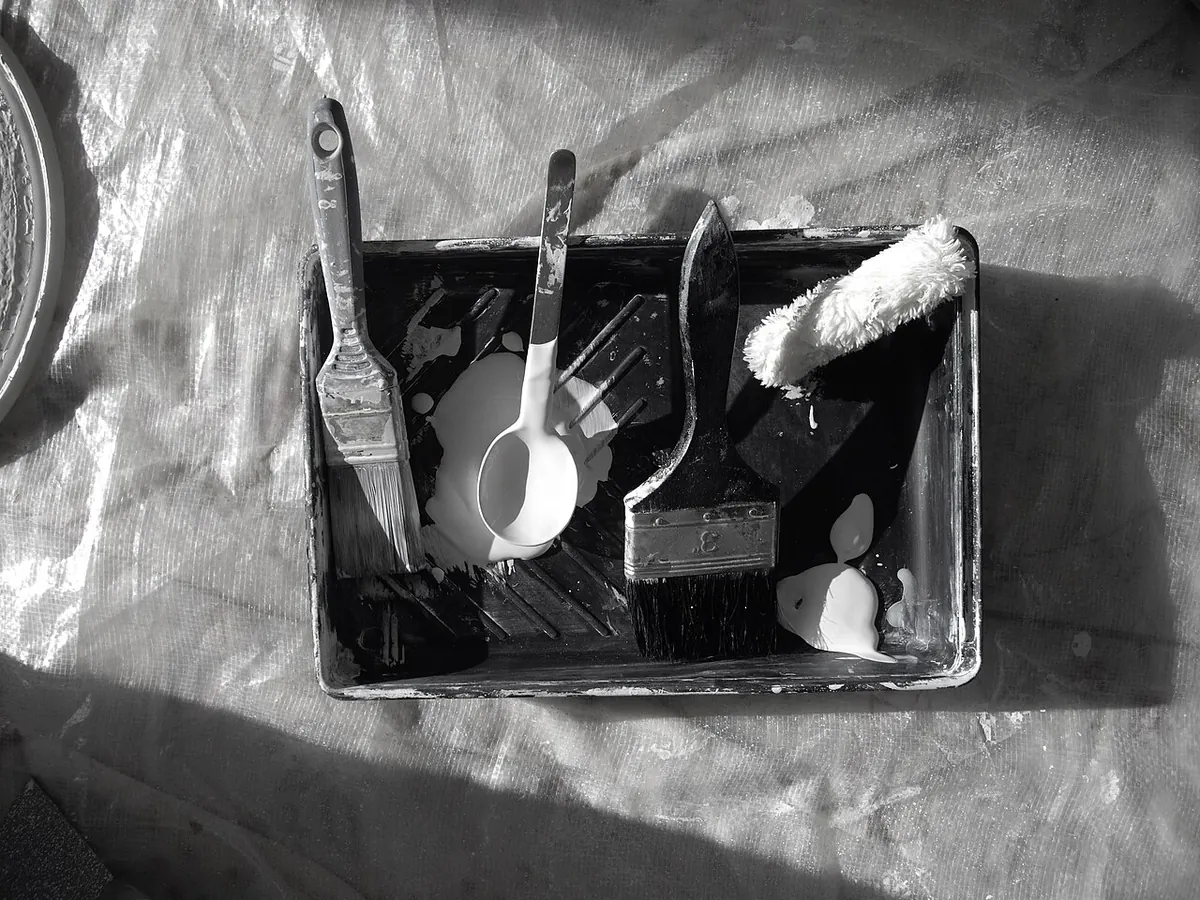 Black and white photo of a paint tray holding brushes, a roller and a paint cup on plastic sheeting