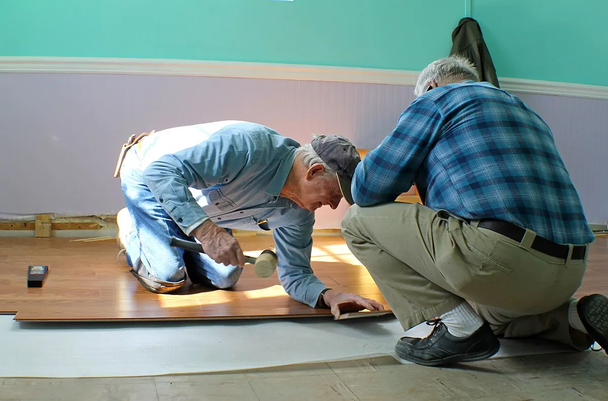 Two installers kneeling and tapping a wood-look laminate plank into place along a wall