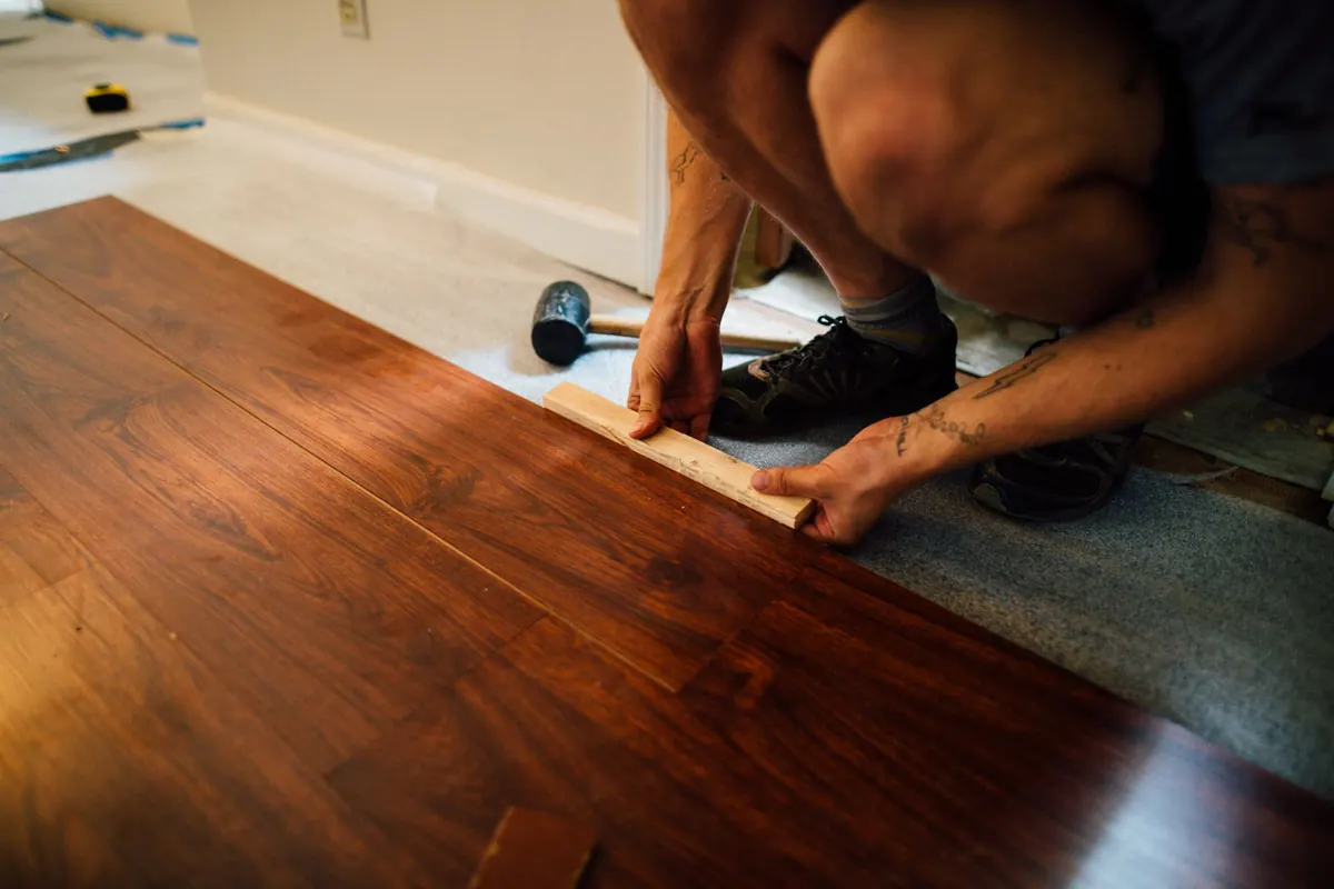 A kneeling installer pressing a dark laminate plank into the previous row with a tapping block, over grey foam underlayment