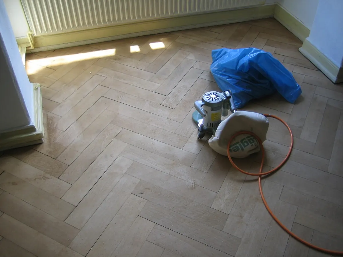 A floor sander at rest on a herringbone parquet floor in an empty room, mid-renovation