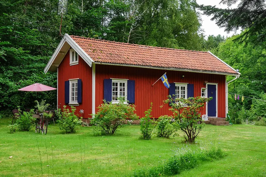 A small red timber cottage with white window frames, dark blue shutters and a tiled roof, standing in a green garden