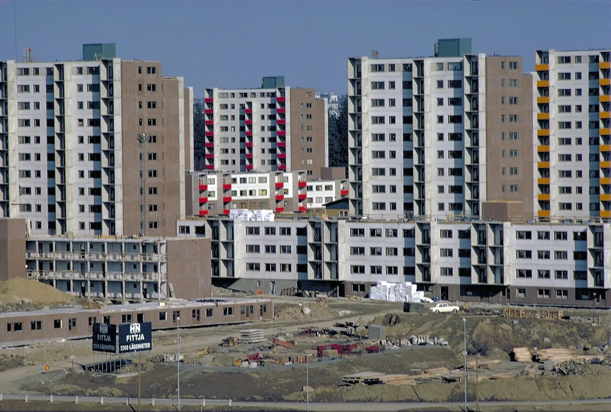 Tall concrete apartment blocks with balconies rising above low buildings on a construction site, with a sign reading Fittja 2200 lägenheter