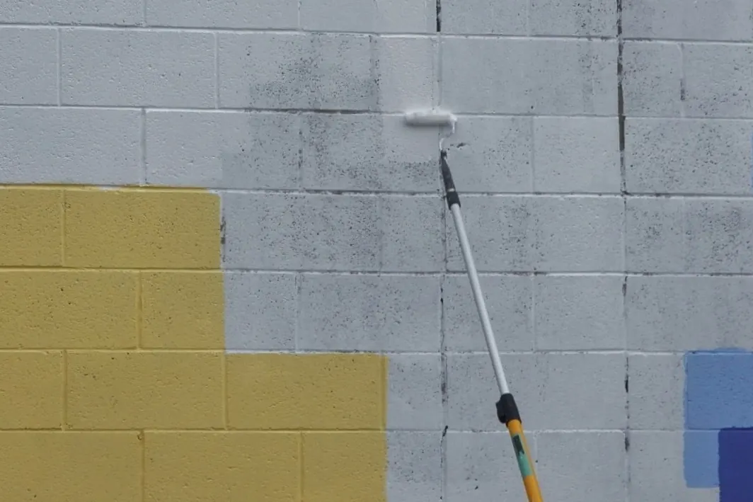 Worker rolling white paint over a yellow block wall, with the first coat still patchy and the old color showing through