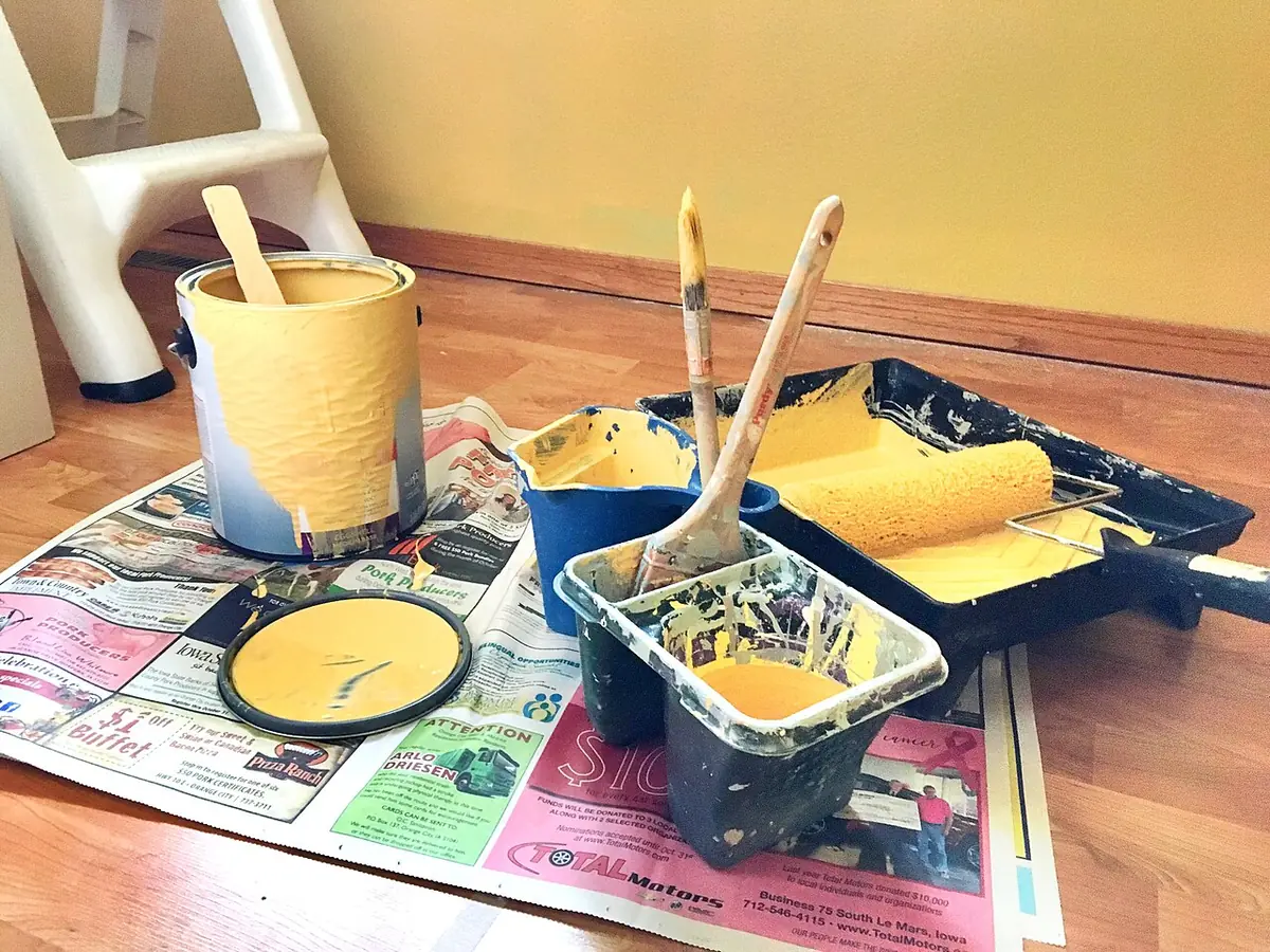 Paint tray with a loaded yellow roller, brushes resting in cups and an open paint can on newspaper