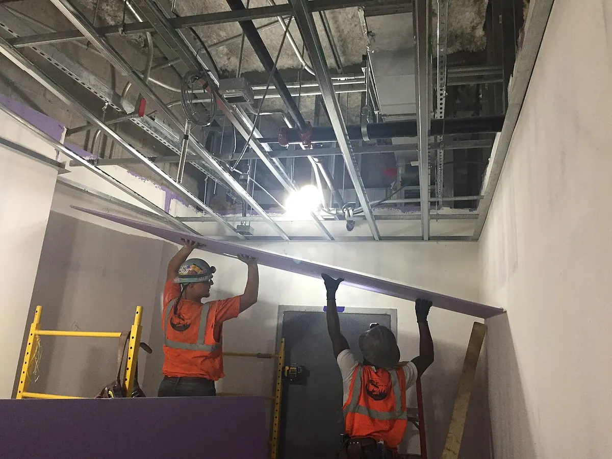 Two workers in hard hats and orange vests lifting a long drywall sheet up to a metal-framed ceiling
