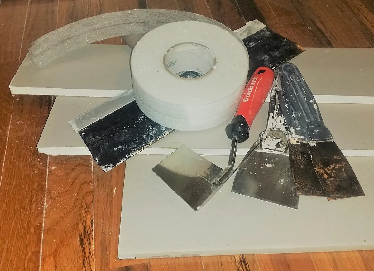 Several pieces of cut drywall on a wooden floor with taping knives and a roll of drywall tape