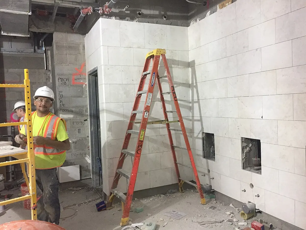 Workers finishing a white tiled wall with cut-outs for plumbing and a ladder in front of it