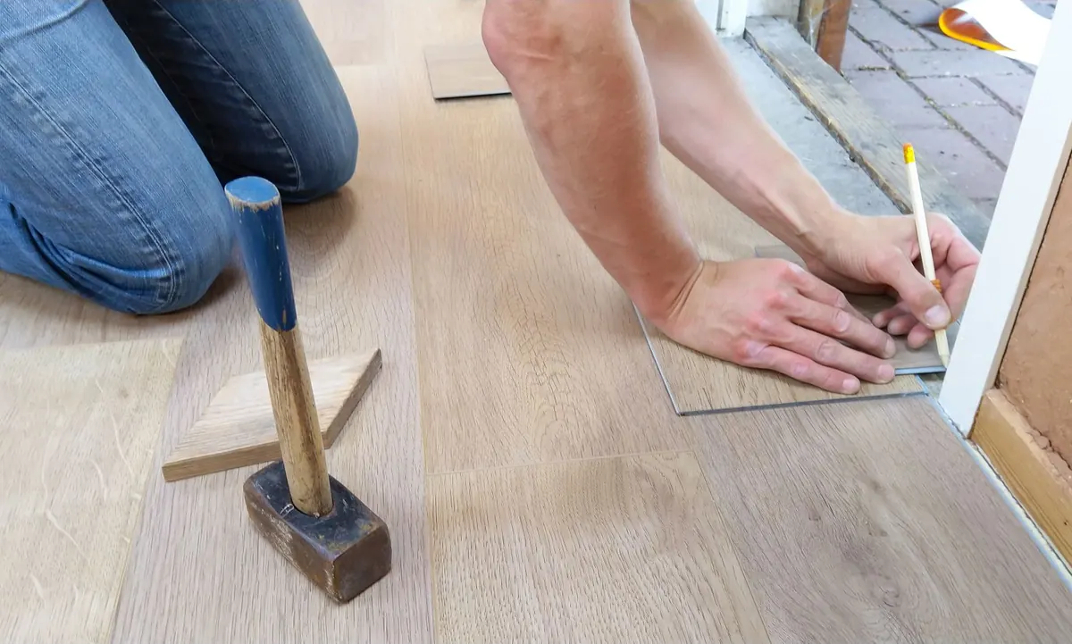 A man marking a cut line with a pencil on a light wood-look plank next to a door frame, with a mallet and tapping block beside him