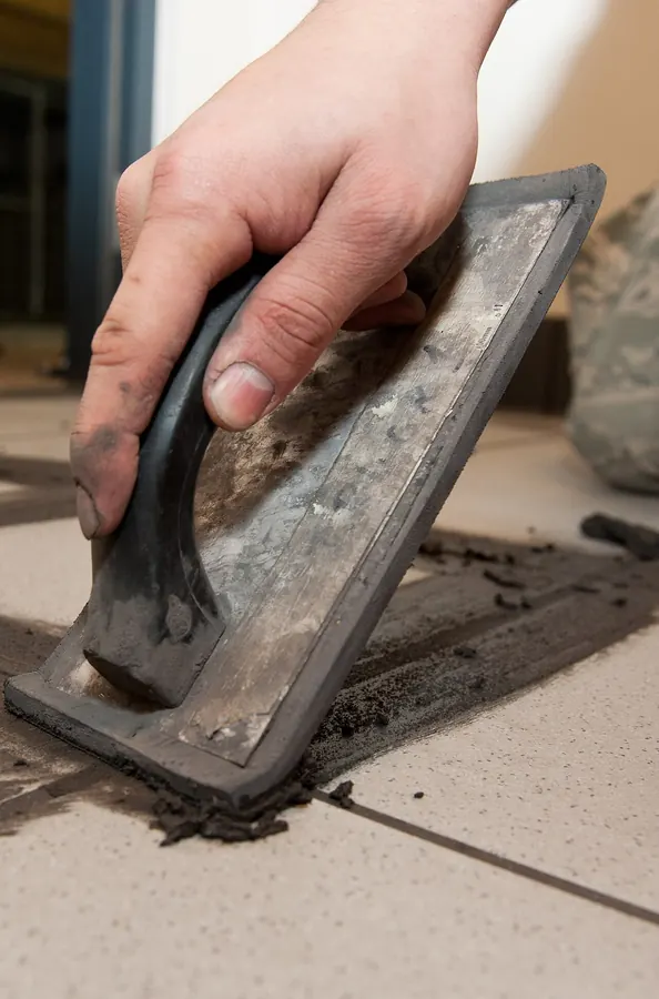 A bare hand pressing a hard rubber grout float across cement grout between large floor tiles