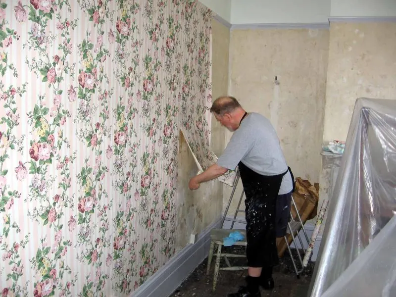 Man smoothing a floral striped wallpaper strip into place beside a bare plaster wall still to be covered