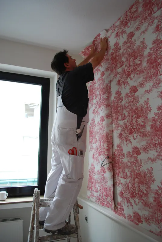 Decorator on a stepladder pressing a red toile wallpaper strip into place at the ceiling line
