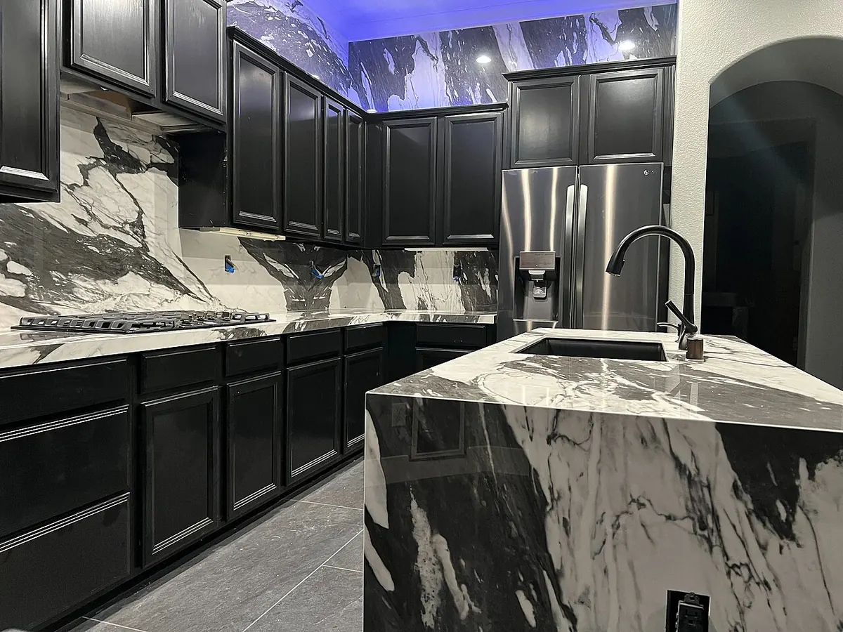 Black cabinets with a white and black veined stone countertop, matching full-height backsplash and a waterfall island with a sink