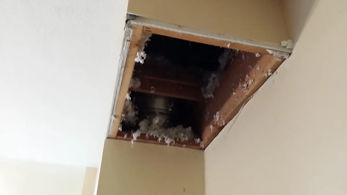 Rectangular opening cut into a ceiling soffit exposing wood framing and loose insulation