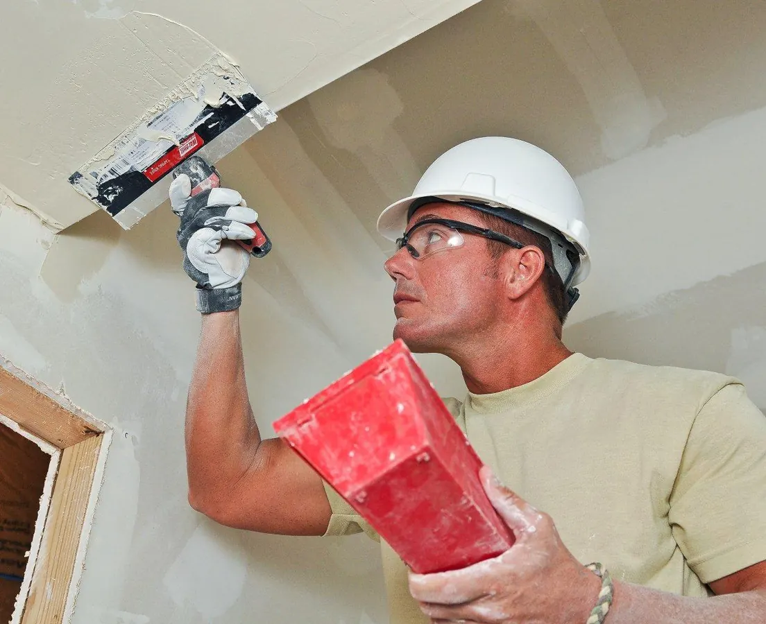 Worker in a hard hat spreading joint compound with a wide taping knife along a drywall seam near the ceiling