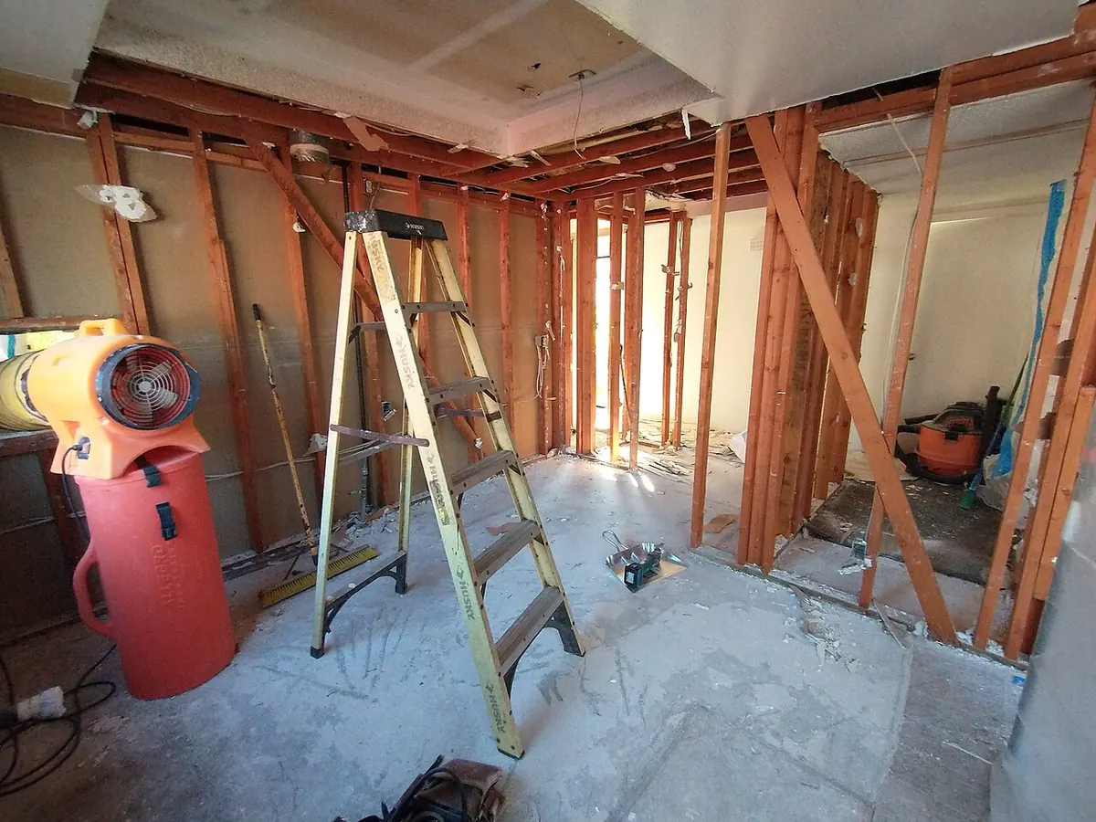 Kitchen stripped to bare wood studs and ceiling joists, with a stepladder, shop vacuum and air mover on a dusty concrete floor