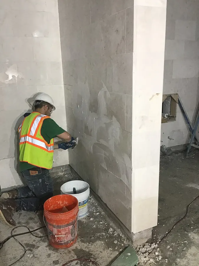 Installer kneeling beside a wall of large pale tiles, spreading grout across the joints with a rubber float
