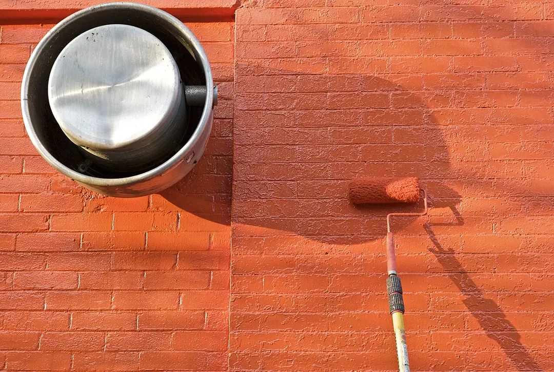 A roller on an extension pole applying orange paint to a brick wall beside a round metal vent