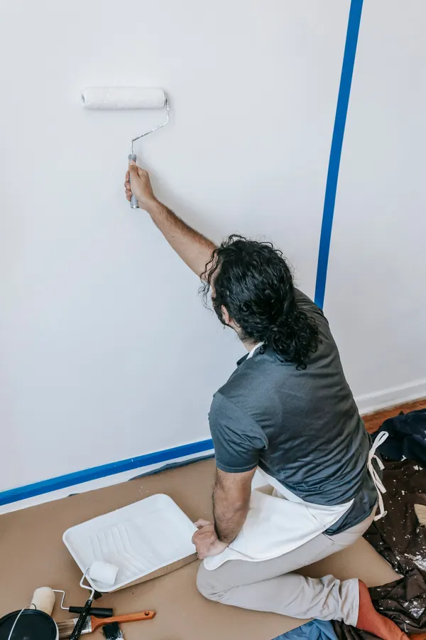 A man kneeling on a drop cloth rolling white latex paint onto a bedroom wall, with blue painter's tape along the corner and a paint tray beside him